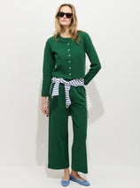 A woman stands against a white background wearing a green buttoned stretch cotton jersey top and matching Coco Sport Pant. She accessorizes with sunglasses, blue flats, and a striped shirt tied around her waist for a chic look.