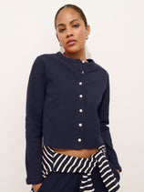A woman stands against a plain background, wearing the Coco Sport Cardigan in navy blue stretch cotton jersey with long sleeves. She has a navy and white striped garment tied at her waist and looks confidently at the camera.