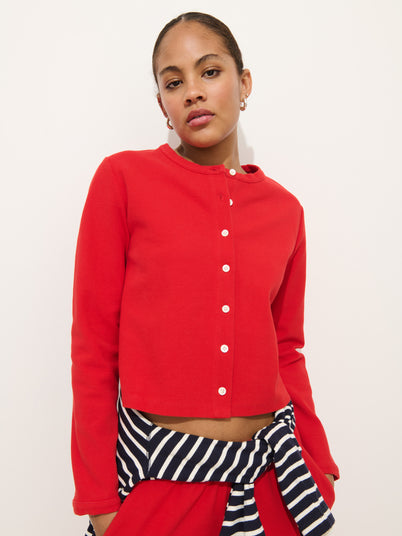 A person wearing women's designer Coco Pants and a bright red Coco Sport Cardigan made from stretch cotton jersey, with a black and white striped sweater tied around their waist, stands against a plain white background.