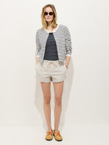 A woman stands against a plain white background with her hands in her pockets, wearing sunglasses and the Coco Track Short In Cotton paired with a striped jacket, striped top, and tan shoes with orange laces.
