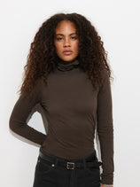 A woman with long curly hair wears the Tissue Turtleneck In Cotton in brown and black jeans, standing against a plain white background with a relaxed expression.