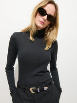 A woman with straight blonde hair wears black cat-eye sunglasses, the Tissue Turtleneck In Cotton, and dark trousers with a black belt; she stands against a plain white background.