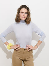 A person with shoulder-length brown hair stands with hands on hips against a white background, wearing the Tissue Turtleneck In Cotton in light blue and tan pants. They accessorize with a yellow scrunchie on one wrist and a silver bracelet on the other.