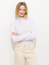 A woman with blonde hair stands against a plain white background, smiling with her arms crossed while wearing a Tissue Turtleneck In Cotton and light beige pants.