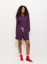 A woman with curly hair stands against a white background, wearing Alex Mill’s The Striped Waffle Short set: a navy blue and red striped long-sleeve shirt, matching drawstring shorts, and red socks with beige toes and heels.