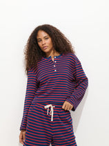 A woman with long curly hair wears The Striped Waffle Sleeping Henley—a navy and red striped pajama set with a buttoned top and drawstring shorts—standing against a plain white background.