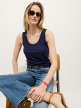 A woman with light brown hair and red bracelets wears sunglasses, blue jeans, brown sandals, and the Garcon Tank In Linen Cotton as she sits on a wooden chair against a plain white background.