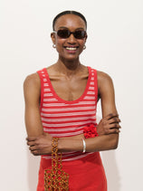 A smiling person in dark sunglasses wears the Garcon Striped Tank In Linen Cotton, a red skirt, and a red scrunchie, holding a beaded bag against a plain white background.