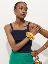 A woman poses against a white background wearing the Lara Tank In Cotton Pointelle, a fitted navy tank with a square neckline, paired with a green skirt. She has a yellow scrunchie on her left wrist, silver bracelet on her right, and holds a brown beaded bag.