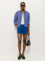 A woman stands against a white background wearing sunglasses, a blue and white striped cardigan over a white top, the Coco Track Short in Cotton, green shoes, and holding a small brown woven handbag.