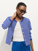A woman in sunglasses smiles, wearing the Coco Sport Striped Cardigan in Cotton over a white top and blue pants, while touching the back of her neck.