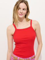 A woman with long, light brown hair wears the Lara Tank in Cotton Pointelle, a sleeveless red square neckline fitted top, paired with pink pants and a tan braided belt, standing against a white background and smiling subtly to her left.