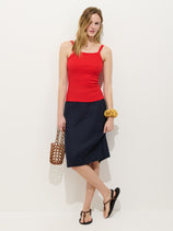 A woman stands against a white wall wearing the Maya Pencil Skirt In Linen with a red sleeveless top, black sandals, a chunky bracelet, and a brown woven handbag. Her wavy hair is styled loosely for an effortlessly chic look.