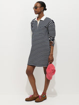Someone stands against a white background wearing the Lizzy Rugby Stripe Dress in cotton jersey, featuring black and white stripes and a feminine silhouette. They wear brown shoes, glasses, and hold a pink cap.