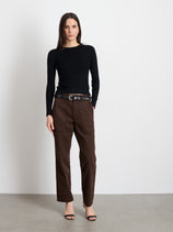 A woman with straight dark hair and a neutral expression stands against a white wall wearing a black long-sleeve top, black open-toe heels, and the Neil Pant in Herringbone—a high-waisted, military-inspired brown trouser with a black belt.