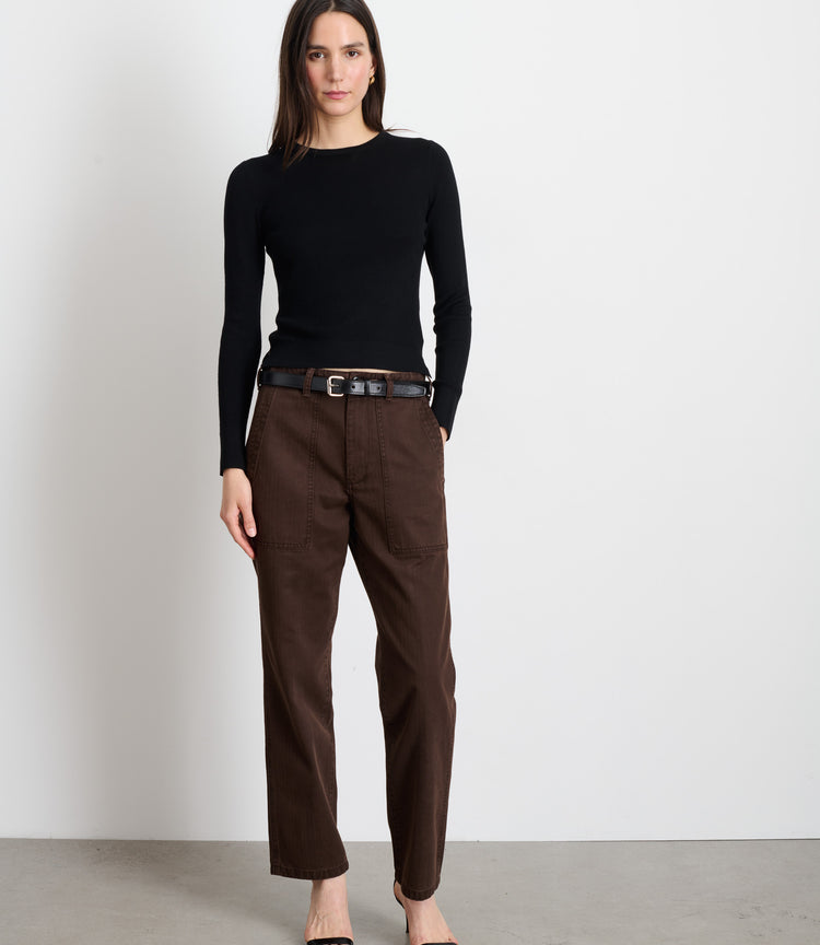 A woman with straight dark hair and a neutral expression stands against a white wall wearing a black long-sleeve top, black open-toe heels, and the Neil Pant in Herringbone—a high-waisted, military-inspired brown trouser with a black belt.