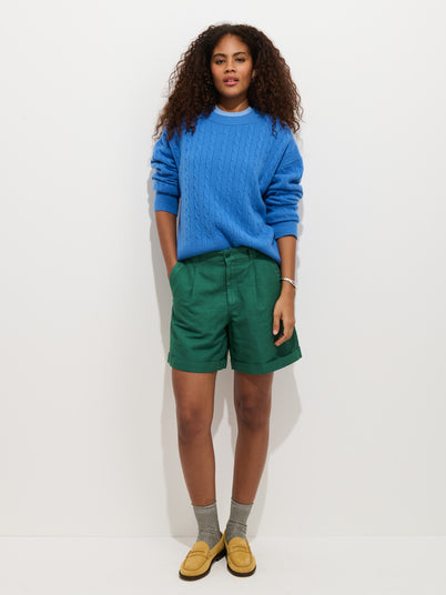 A woman with curly hair stands against a white background wearing a blue cable-knit sweater, gray socks, yellow loafers, and Lucie Short In Linen Cotton—green shorts with an elastic waistband. She looks relaxed with her hands in her pockets.