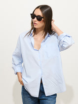 Wearing the Jo Shirt In Cotton Poplin, an oversized light blue 100% cotton shirt, with blue jeans and black sunglasses, a person stands against a white background, looking to the side and touching their hair.