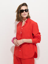 A person with shoulder-length brown hair wears black sunglasses and the Jo Shirt in Cotton Poplin—a bright red, 100% cotton button-up featuring Troca shell buttons—paired with matching pants, standing against a plain white background. They accessorize with a pink scrunchie and a silver bracelet on their left wrist.
