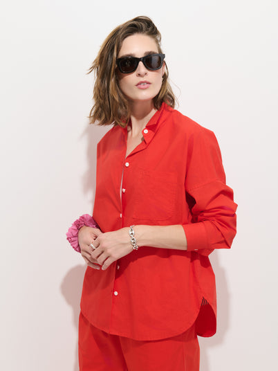 A person with shoulder-length brown hair wears black sunglasses and the Jo Shirt in Cotton Poplin—a bright red, 100% cotton button-up featuring Troca shell buttons—paired with matching pants, standing against a plain white background. They accessorize with a pink scrunchie and a silver bracelet on their left wrist.