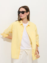 A woman wears black sunglasses, white pants, and the Jo Shirt In Cotton Poplin—a yellow 100% cotton button-down—layered over a white T-shirt, posing with one hand on her hip against a plain light background.