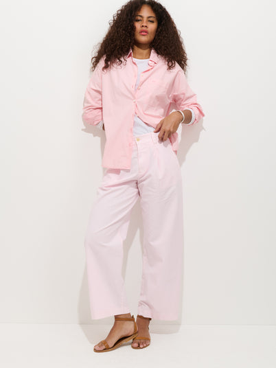 A woman with curly hair poses against a white background, wearing the Soho Pleated Pant In Cotton Linen styled with a light pink button-up over a white top and tan sandals. She stands with one hand in her pocket and looks relaxed.
