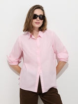 A woman stands against a white background with her hands in her pockets and sleeves rolled up, wearing the Jo Shirt In Cotton Poplin—a light pink 100% cotton button-up featuring Troca shell buttons—and dark sunglasses.