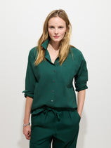 A woman with long, blonde hair stands against a white background, wearing the Jo Shirt in Cotton Poplin—a dark green 100% cotton button-up shirt with matching pants and sleeves rolled up. Troca shell buttons provide a subtle accent to her look as she faces the camera with a neutral expression.