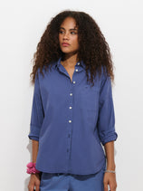 A woman with curly hair wears the Jo Shirt in Cotton Poplin, a loose 100% cotton blue button-up featuring French seams and rolled sleeves, paired with blue pants, a pink scrunchie on her wrist, and a silver bracelet, standing against a plain white background.