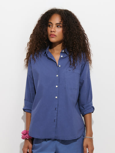 A woman with curly hair wears the Jo Shirt in Cotton Poplin, a loose 100% cotton blue button-up featuring French seams and rolled sleeves, paired with blue pants, a pink scrunchie on her wrist, and a silver bracelet, standing against a plain white background.