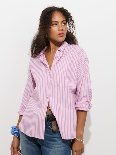A woman with curly hair stands against a white background, wearing the Jo Shirt In Stripe Cotton Poplin—a loose-fitting, light pink striped 100% cotton button-up—paired with blue jeans. She looks to the side with one hand in her jeans pocket and a blue scrunchie on her wrist.