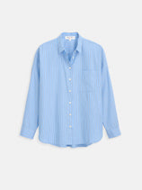 The Jo Striped Shirt In Cotton Poplin features an oversized light blue design with long sleeves, thin vertical blue and red stripes, a collar, white buttons, and a single chest pocket, shown against a plain white background.