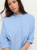 A woman with long brown hair wears the Jo Striped Shirt in Cotton Poplin—an oversized light blue shirt with white and red stripes—standing against a plain white background and looking at the camera.