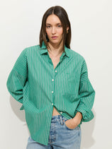 A woman with straight brown hair wears the Jo Striped Shirt In Cotton Poplin—a loose, green and white striped 100% cotton button-up with Troca shell buttons—paired with light blue jeans, standing against a plain white background with one hand in her pocket.