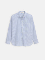 The Jo Striped Shirt In Cotton Poplin, featuring blue and white vertical stripes, long sleeves, front pocket, collar, and French seams, is crafted from 100% cotton and shown laid flat on a plain white background.