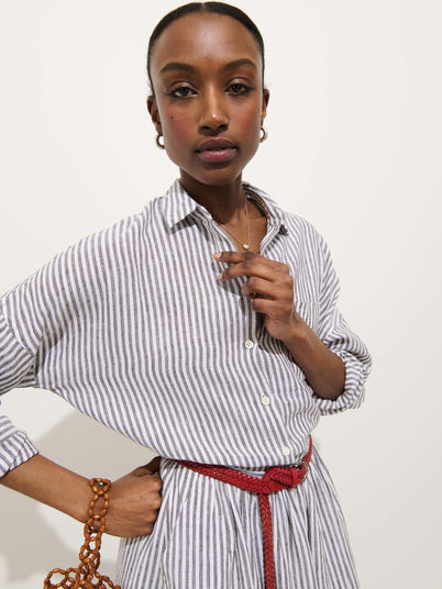 Someone stands against a white background wearing the Jo Striped Shirt In Linen, a relaxed fit, long-sleeved, white and gray striped 100% linen dress with real shell buttons and a red braided belt, holding a brown beaded bag.