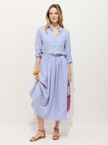 A woman stands against a plain background wearing the Sarah Striped Skirt In Linen with a matching blue and white striped linen button-down shirt, tan sandals, a pink purse, and a yellow scrunchie on her wrist.