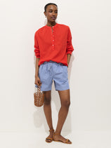A person stands against a white background wearing a bright red long-sleeve shirt, Hudson Striped Short In Poplin, brown sandals, and holding a small woven bag.
