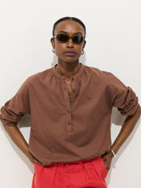 A person wears sunglasses, red pants, and the Nina Striped Popover in Cotton Voile—a relaxed-fit brown women’s long-sleeve button shirt—standing against a plain white background with arms relaxed by their sides.