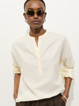 A person wears the Nina Striped Popover In Cotton Voile with rolled-up sleeves, sunglasses, and dark pants, standing against a plain white background with hands in pockets.