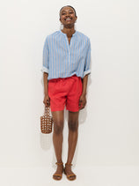A woman stands smiling against a plain white background, wearing the Hudson Short In Linen—red elastic waist summer shorts—paired with a light blue striped button-up shirt, tan sandals, and holding a small woven brown handbag.