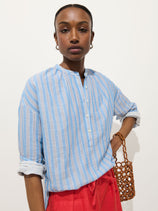 A woman wearing the Nina Striped Popover in Double Gauze—an oversized women's shirt in light blue and white stripes with rolled-up sleeves—pairs it with bright red pants and holds a brown woven bag against a plain white background.