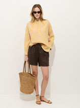 A woman stands against a white wall wearing a yellow striped button-up, Hudson Short In Linen with an elastic waist, flat sandals, sunglasses, and holding a woven straw tote bag.