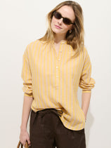A person stands against a plain light background wearing sunglasses, brown pants, holding a woven bag, and the Nina Striped Popover In Double Gauze—a women's oversized fit shirt in yellow and white striped 100% cotton gauze.