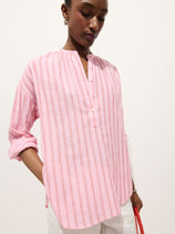 Wearing the Nina Striped Popover In Double Gauze—a light pink, oversized women's blouse with red stripes and rolled-up sleeves—a person stands holding a red item against a plain background.