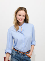 A woman with light brown hair, wearing the Chantilly Ruffle Shirt In Cotton Voile and jeans with a brown belt, stands against a plain white background holding a red bag and smiling slightly at the camera.