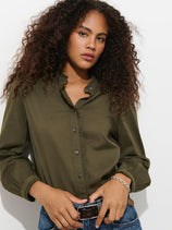 A woman with long curly hair wears the Chantilly Ruffle Shirt in Cotton Voile—a French-inspired olive green button-up crafted from 100% cotton and finished with Troca shell buttons—paired with blue jeans. She stands against a white background, hands resting on her belt and her expression neutral.