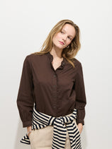A woman with light brown hair wears the Chantilly Ruffle Shirt in Cotton Voile and light pants, standing against a white background with a striped sweater tied at her waist, embodying effortless French girl style as she gazes to the side.