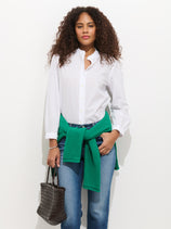A woman with curly hair wears the Chantilly Ruffle Shirt in Cotton Voile, jeans, and a green sweater tied at her waist, standing against a white background and holding a textured black handbag in her left hand.