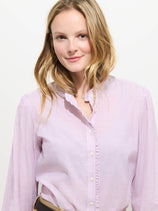 A woman with long blonde hair wears the Chantilly Striped Ruffle Shirt in Cotton Voile, a French-inspired blouse with light pink and white stripes and delicate Troca shell buttons. She smiles softly at the camera against a plain background.